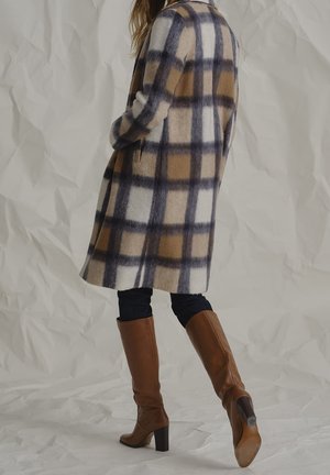 Vrouw draagt een knielange, geruite jas in bruin, beige en zwart, zwarte broek en bruine leren laarzen met hakken, lopend tegen een neutrale achtergrond.