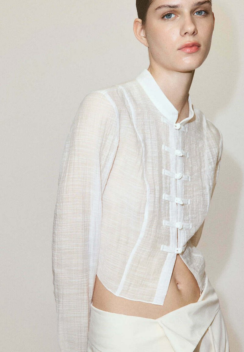 Jeune femme aux yeux bleus porte un chemisier blanc transparent boutonné et un pantalon clair taille haute sur un fond neutre uni.