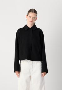 Zwarte cropped jasje met een kraag, lange mouwen en een gladde textuur. Gepaard met witte broek, wat een kleurcontrast creëert.