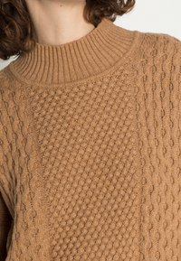 Brun stickad tröja med hög ribbad krage, texturerade mönster och en blandning av bikakestruktur och flätstickning.
