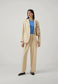 Beige tailored suit with a single button blazer and wide-leg trousers, worn over a blue fitted top. Black flat shoes complete the outfit.