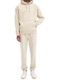 Sudadera beige con un bolsillo canguro y capucha con cordón, combinada con pantalones de chándal ajustados a juego. El material parece suave y liso.