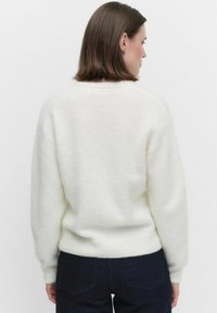 Vrouw met kort bruin haar, gekleed in een witte fluffy trui en donkere jeans, gezien van achteren tegen een effen achtergrond.