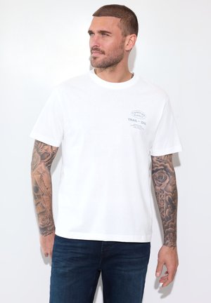 Mann mit kurzen Haaren und Ärmeltattoos trägt ein weißes T-Shirt mit kleinem Logo auf der Brust und dunkelblaue Jeans, blickt nach links.