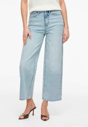 Frau trägt hellblaue Jeans mit weitem Bein, weißes ärmelloses Oberteil und braune Sandalen mit Absatz, steht mit entspannten Händen.