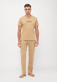 Beige pamut póló, rajta a "POLO RALPH LAUREN" logóval, párosítva mintás bézs nadrággal, amelyen kis sötét ikonok találhatók, könnyű kialakítással.