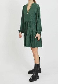 Grön, långärmad klänning med lagerdesign, v-tyg och ögonmönster. Bärs med svarta ankelstövlar på en modell som står.