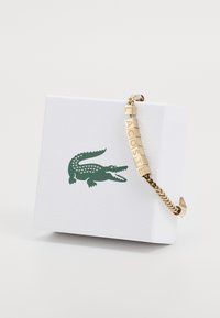 Bracelet en or avec des accents de lettres épelant "LACOSTE", présenté sur un emballage texturé blanc avec un logo de crocodile vert.