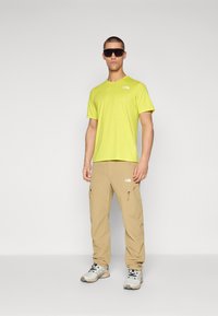 Camiseta de manga corta amarilla brillante con un pequeño logo. Pantalones cargo color beige con bolsillos con cremallera. Botas de senderismo grises y negras. Gafas de sol negras.