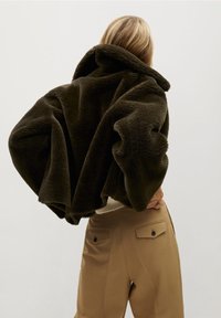 Mørkebrun oversized fleecejakke med høj krave, blød tekstur og voluminøse ærmer, båret over en lys sweater og beige bukser.