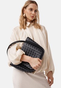 Sac à main en cuir tissé noir avec une forme structurée et une seule poignée, tenu par une personne portant un blouse couleur crème. Détails de la surface texturée visibles.