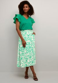 Zelený top s výstrihom do V a volánovými rukávmi doplnený dlhou sukňou s kvetinovým vzorom v zelenej a bielej farbe. Čierne sandále na remienky zvýrazňujú outfit.
