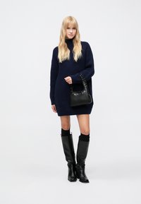 Jūras zila adīta pārmērīga izmēra džemperkleita ar augstu apkakli, kopā ar melnām ceļgalu augstām zābakām un melnu teksturētu rokassomu ar sudraba akcentiem.