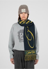 Kobieta w szarej czapce beanie, szarym swetrze z emblematem, ciemnoszarych spodniach oraz granatowo-żółtym szalu z frędzlami owiniętym wokół szyi.