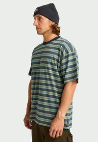Homem em um gorro azul-marinho, vestindo uma camisa de manga curta solta com riscas verdes e azuis, posando de perfil contra um fundo simples.