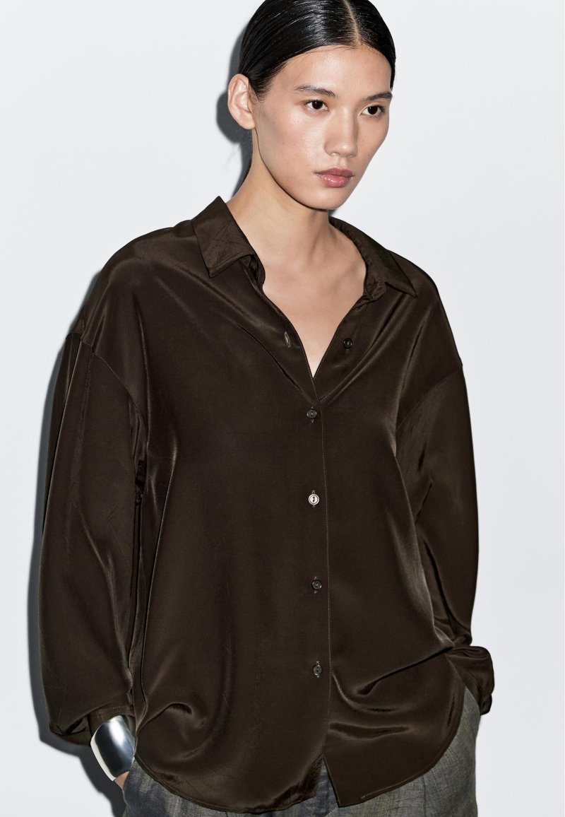 Mørkebrun satinbluse med afslappet pasform, som har knaplukning foran og lange ærmer. Glat tekstur med en diskret glans. Sølvdetalje ved manchet.
