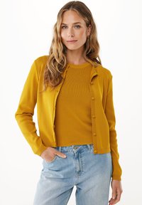 Blondýnka s dlouhými vlnitými vlasy, oblečená v hořčicově žlutém kardiganu přes odpovídající top a světle modré džíny s vysokým pasem, ruka v kapse.