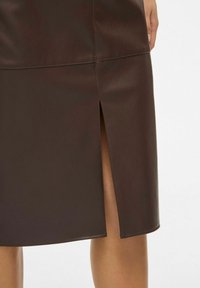 Donkere brown leren rok met een split aan de achterkant, gedragen door een persoon met zichtbare onderbenen en een hand rustend bij de heup.