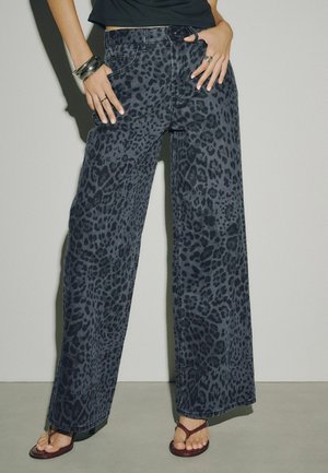 Femme portant un pantalon large à imprimé léopard foncé, un top court noir, des bracelets en argent, des bagues et des sandales marron, debout contre un mur uni.