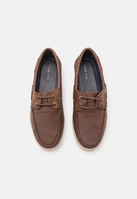 Mocasines de cuero marrón con cordones decorativos, costuras claras y una superficie texturizada. Presentan una suela de goma beige lisa.