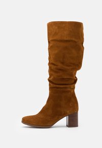 Brązowy zamszowy trzewik do kolan o luźnym fasonie, kwadratowym nosku i grubym, fakturowanym obcasie w kształcie słupka. Posiada gładką cholewkę i minimalistyczny styl.