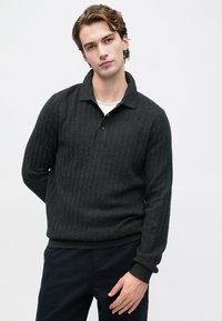 Jovem com cabelo ondulado vestido com um suéter polo de manga longa cinza escuro texturizado por cima de uma camiseta branca, posando contra um fundo claro e simples.