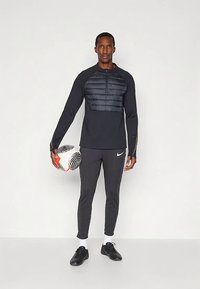 Férfi sportoló fekete sportöltözetben egy focilabdát tart, áll egy sima fehér háttér előtt.