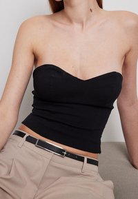 Top nero senza spalline con scollatura a cuore, corpino aderente e texture liscia, abbinato a pantaloni beige e una cintura nera.