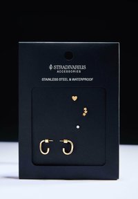 Sæt af øreringe i guldtonet rustfrit stål med åbent hoop-design, små hjerte-, stjerne- og mistelten-accents på et sort bagkort.