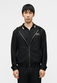 Μαύρο αθλητικό μπουφάν με υφή στο ύφασμα, φερμουάρ και κουκούλα. Διαθέτει λογότυπο και χρυσές λεπτομέρειες στο γιακά και τις ραφές.