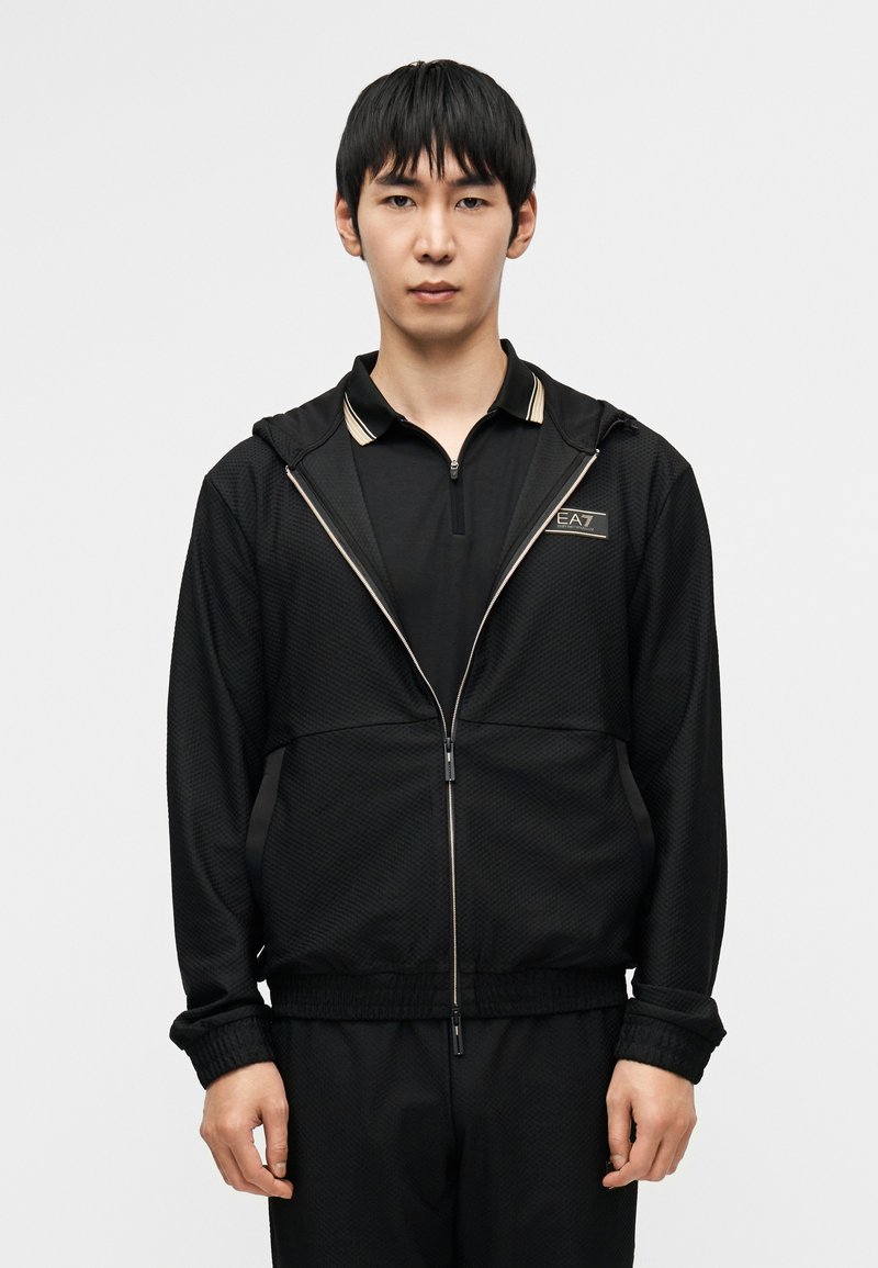 Μαύρο αθλητικό μπουφάν με υφή  στο ύφασμα, φερμουάρ και κουκούλα. Διαθέτει λογότυπο και χρυσές λεπτομέρειες στο γιακά και τις ραφές.