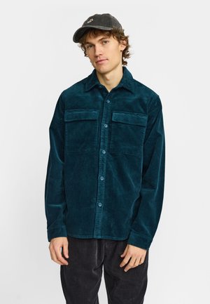 Man draagt een tealcorduroy blouse met knopen en zwarte pet, staat met ontspannen handen tegen een effen witte achtergrond.