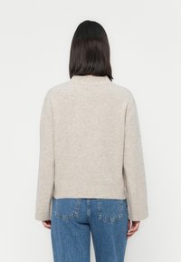 Beige pulóver bordázott alsó résszel és mandzsettákkal, puha anyagból készült, kerek nyakkivágással és kissé laza szabással. Kék farmernadrággal viselve.