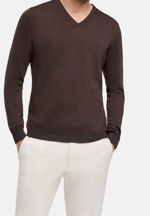 Man wearing a fitted dark brown V-neck long-sleeve sweater with hands in pockets, paired with off-white trousers.