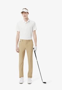 Vêtements de golf comprenant une chemise polo blanche avec col, un pantalon beige, des chaussures blanches et une visière ; tenant un club de golf avec une main gantée.