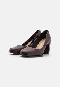 Zapatos de tacón alto de cuero marrón con punta curva y tacón grueso. El interior cuenta con una plantilla perforada para una mayor transpirabilidad.