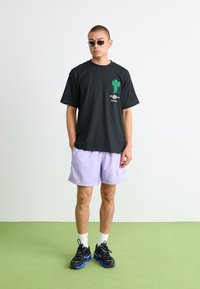 Sort t-shirt med et grafisk motiv af en grøn kaktus og tekst, parret med lavendelfarvede shorts. Modellen bærer sorte sneakers og smalle solbriller.