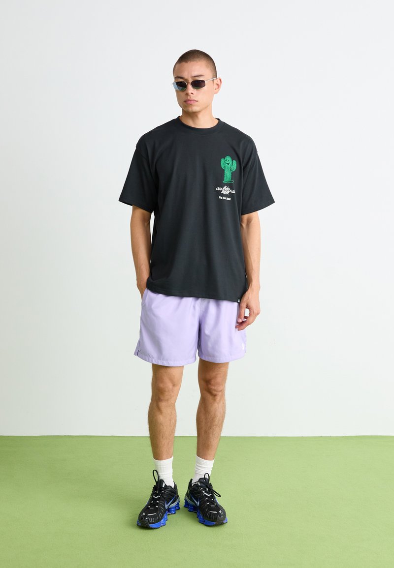 T-shirt noir avec un graphique de cactus vert et du texte, associé à un short lavande. Le mannequin porte des baskets noires et des lunettes de soleil étroites.