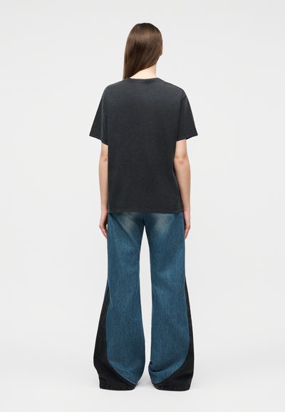 Άτομο με μακριά καστανά μαλλιά που φοράει μαύρο κοντομάνικο μπλουζάκι και φαρδιά μπλε τζιν παντελόνια με μαύρες πλευρικές ραφές, όρθιο με την πλάτη προς τα εμπρός.