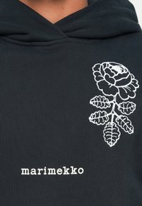 Melna kapuce ar ribotu tekstūru, kurai ir balta rozes grafika un zīmola nosaukums "marimekko", kas izdrukāts baltā krāsā zem dizaina.