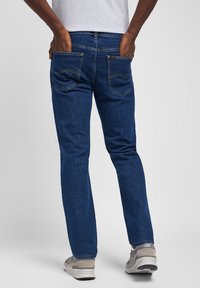 Jeans de hombre de mezclilla azul con corte recto, con bolsillos traseros y detalles de costura amarilla. El tejido parece suave y duradero.