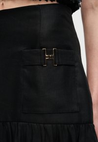 Črna krilo z žepom spredaj, na katerem je zlata kovinska zaponka v obliki črke H, in nabranim detajlom na robu.