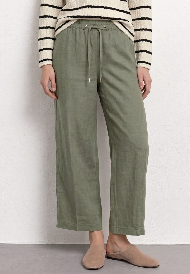 Pantalon ample vert olive clair avec taille à cordon, porté avec des chaussures beiges à enfiler et un pull rayé noir et blanc.