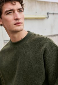 Olivgrüner, strukturierten Fleece-Pullover mit einem gerippten Kragen, der einen lockeren Schnitt und sichtbare Steppnähte aufweist.