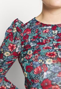 Bluse med blomsterprint og krøllet tekstur; har puffede ærmer og en rund halsudskæring. Dominerende farver inkluderer rød, blå og grøn.