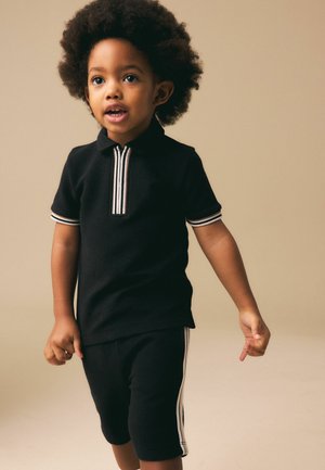 Kleinkind mit lockigem Haar, das ein schwarzes, kurzärmeliges Polo mit Reißverschluss und passende Shorts mit weißen Streifen trägt, vor beigem Hintergrund stehend.