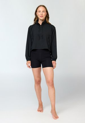 Mujer joven de pie descalza con una sudadera con capucha corta negra de manga larga y pantalones cortos ajustados negros sobre un fondo claro y liso.