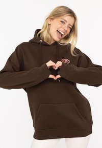 Brązowa bluza z kapturem, z kieszenią z przodu, sznurkiem do regulacji, oraz różowym haftem w kształcie serca. Miękka tkanina i luźny krój z długimi rękawami.