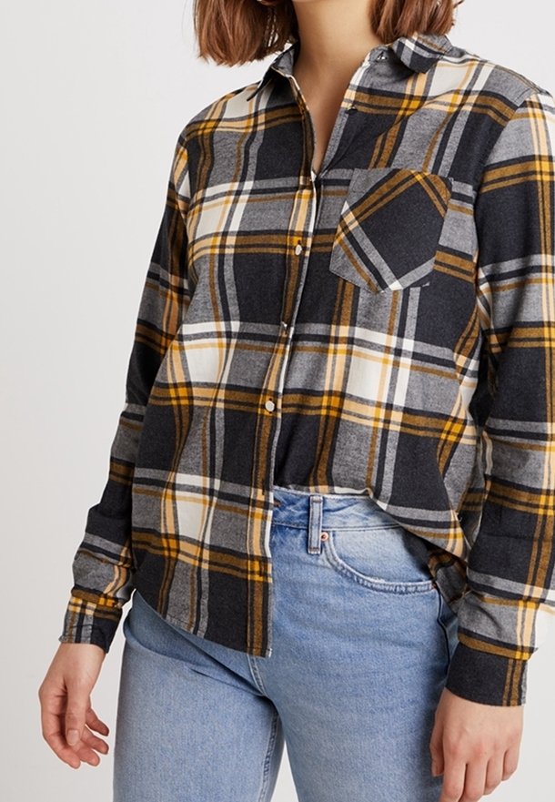 Femme portant une chemise à manches longues à carreaux noirs, gris, blanc et jaune, avec un jean bleu clair.
