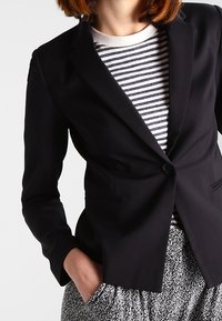 Frau in einem taillierten schwarzen Blazer über einem gestreiften Hemd, mit ihrer Hand in der Tasche einer gemusterten grau-weißen Hose.
